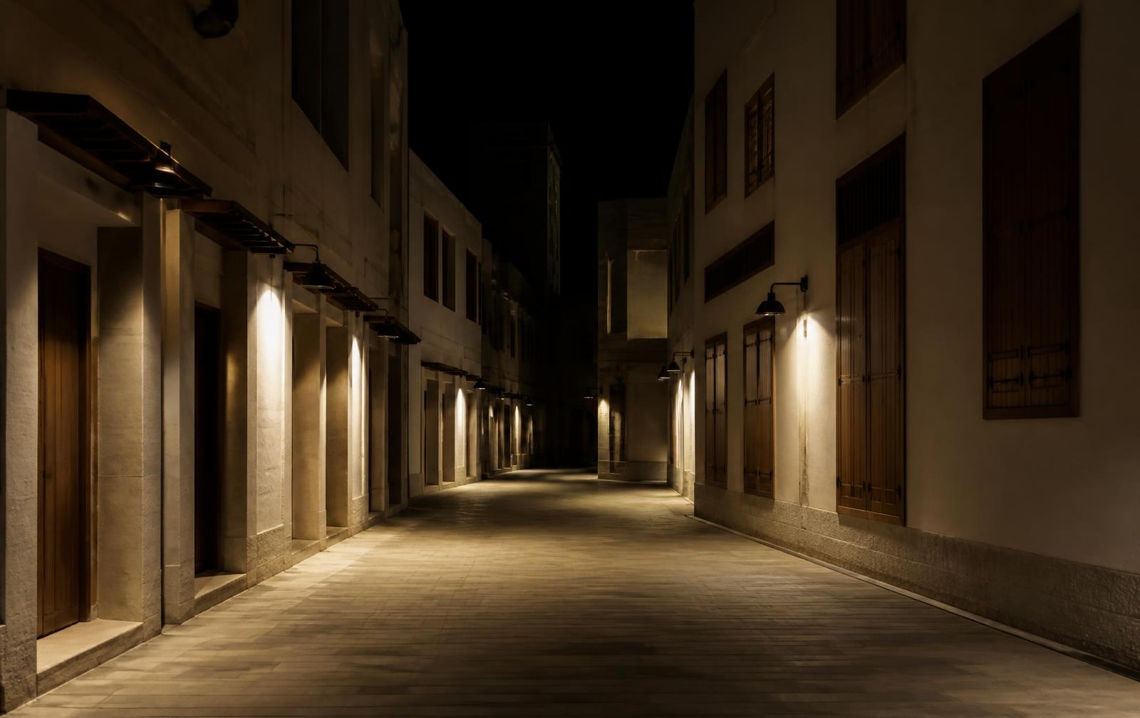 A narrow empty pedestrian street at night between pale stone buildings with wooden shutters, warm lamplight pooling on the paving