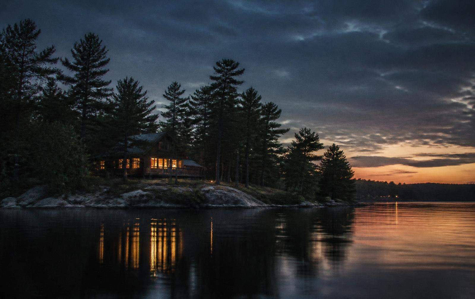 Muskoka lake at dusk with a dark boathouse, warm lit windows and still water reflecting pines