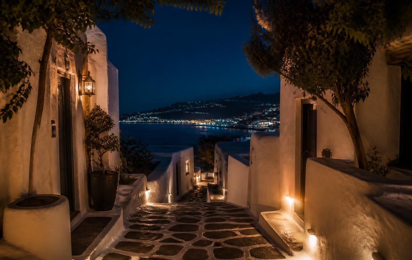 A lantern-lit whitewashed lane in Mykonos at night with Aegean lights beyond