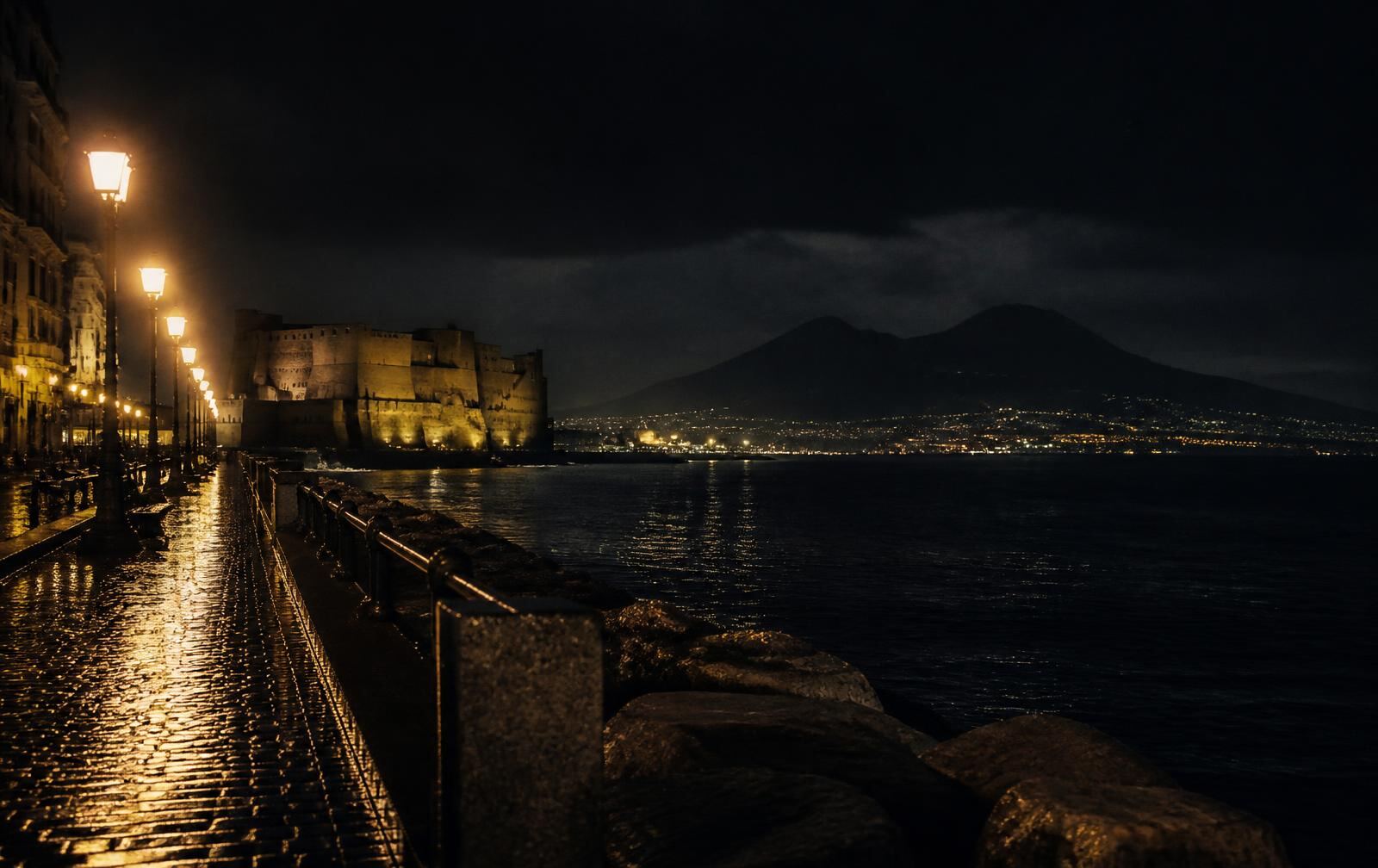 The Naples seafront at night with lamplit promenade, a floodlit seaside castle and Vesuvius across the dark bay