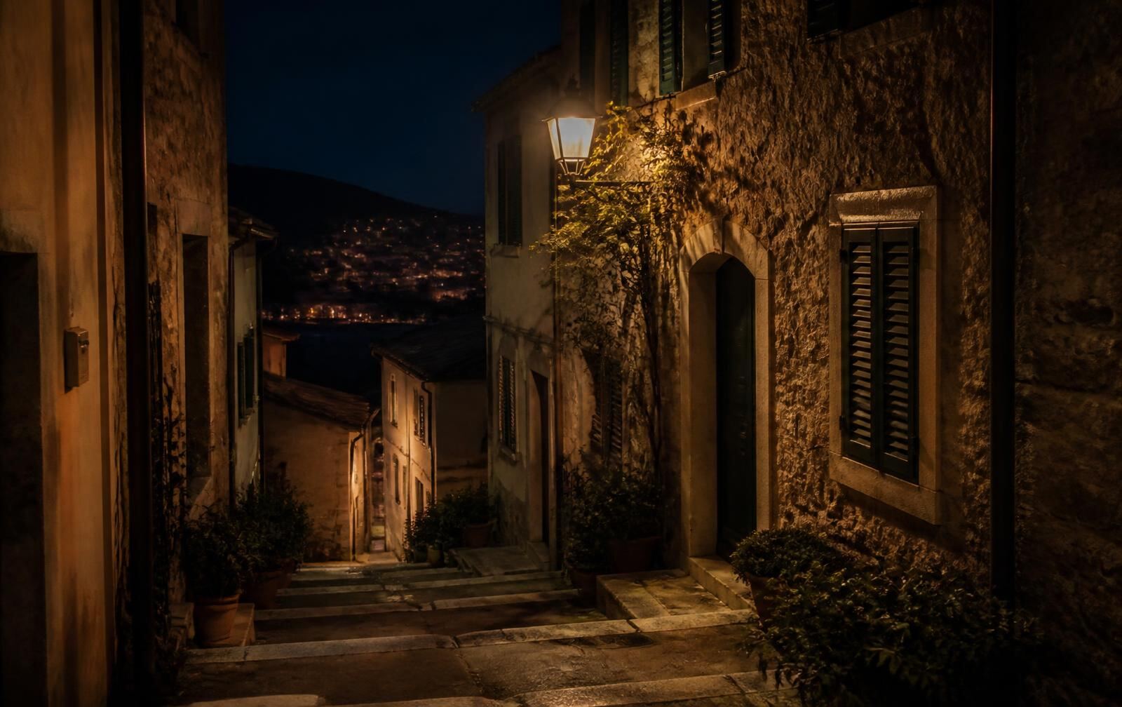 Narrow stepped hillside street at night with pale stone walls, shuttered windows and a single warm lamp
