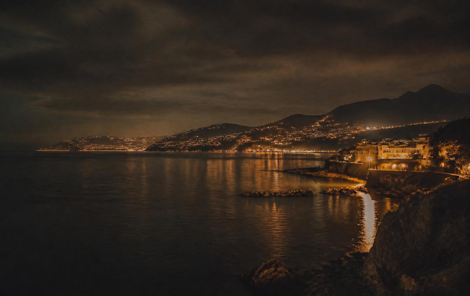 A dark coastline and harbour at night with distant city glow reflected on black water