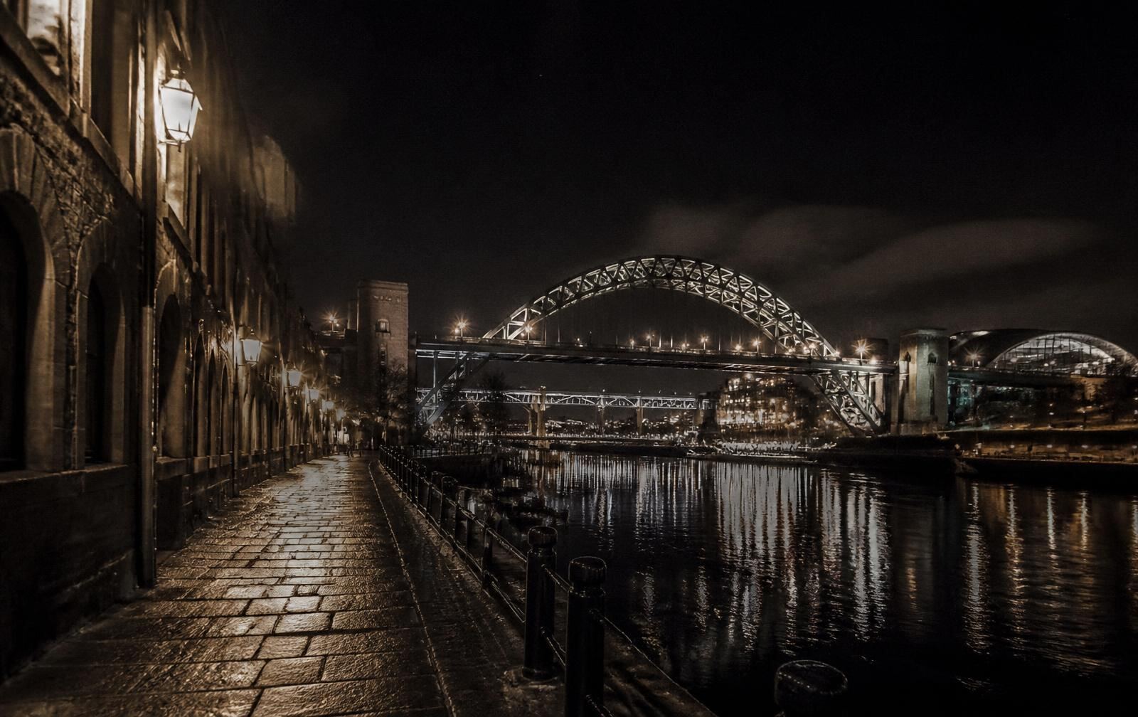 Newcastle quayside at night with lit bridge arch reflected in the dark River Tyne