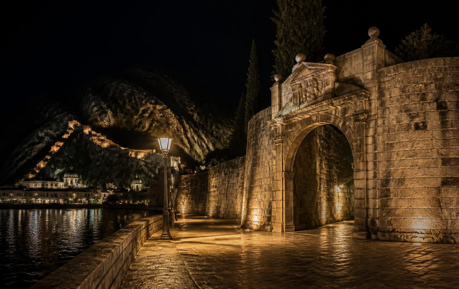 An old stone fortress gate and riverside promenade lit warm gold at night in a southern Balkan city