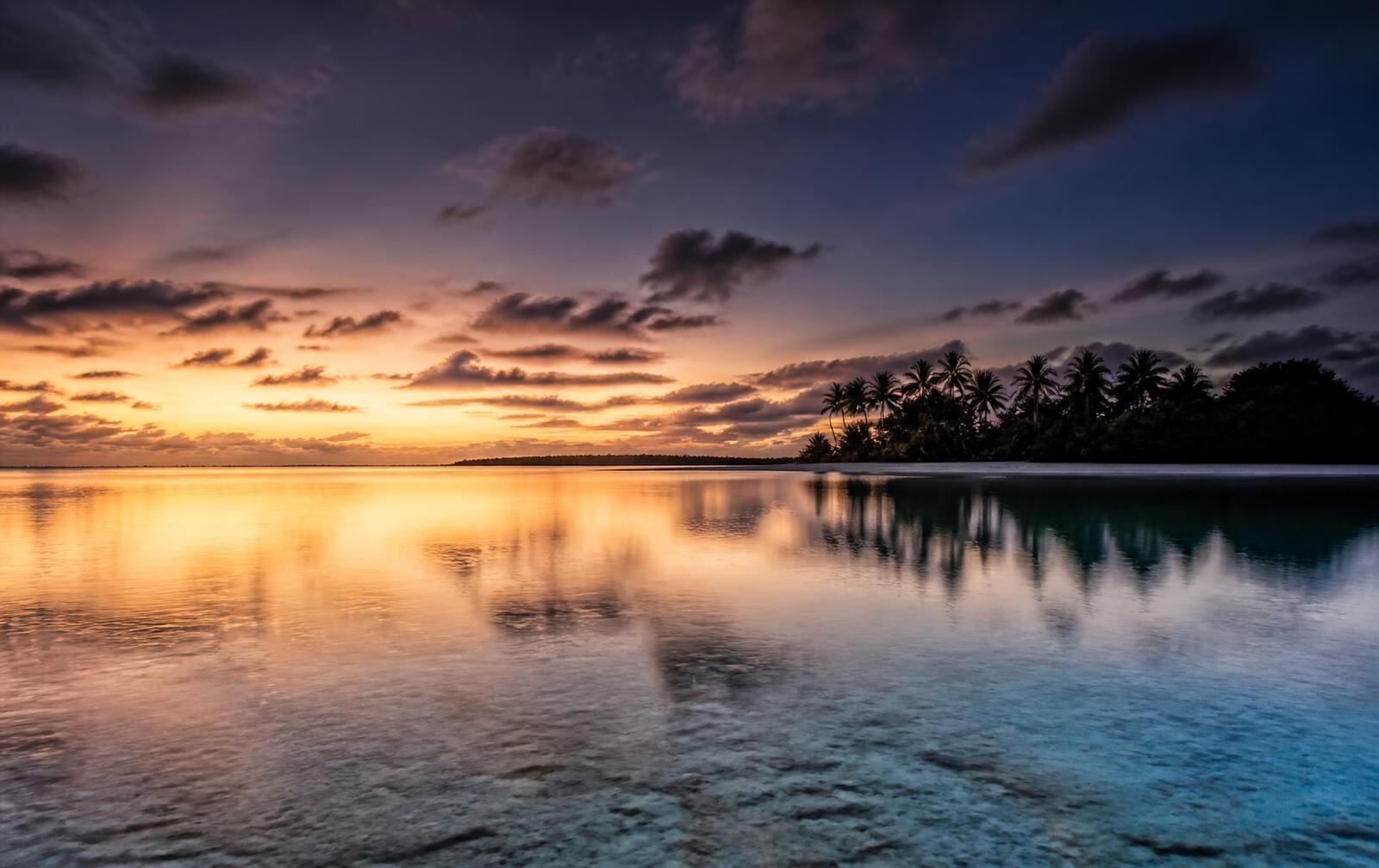A wide flat lagoon at dusk reflecting pale gold light with a low palm island on the horizon