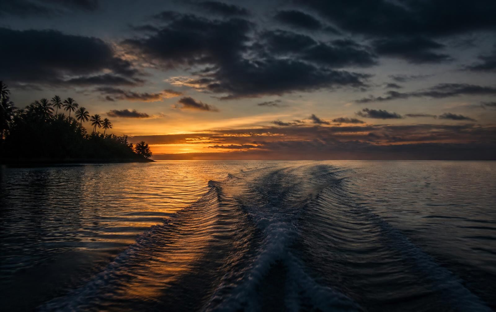 A boat wake fading across a calm lagoon at sunset with a palm island silhouetted against gold light