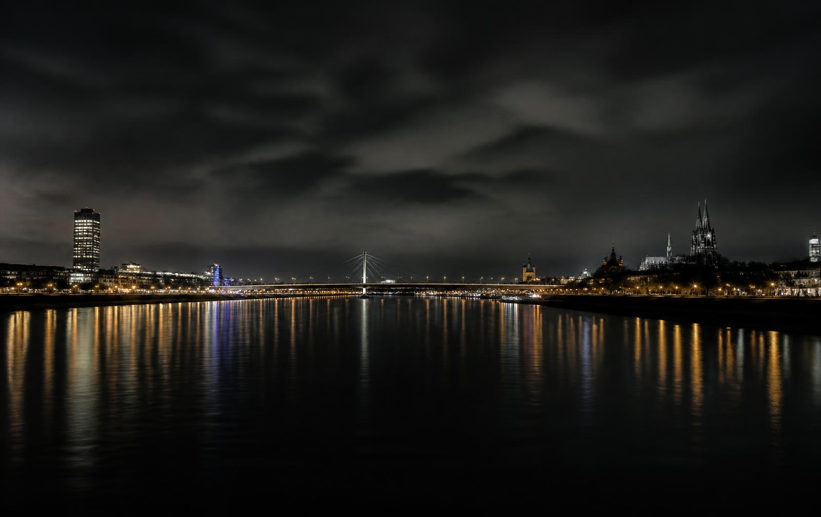 The Rhine at night with city lights and a bridge reflected across black water under a heavy sky