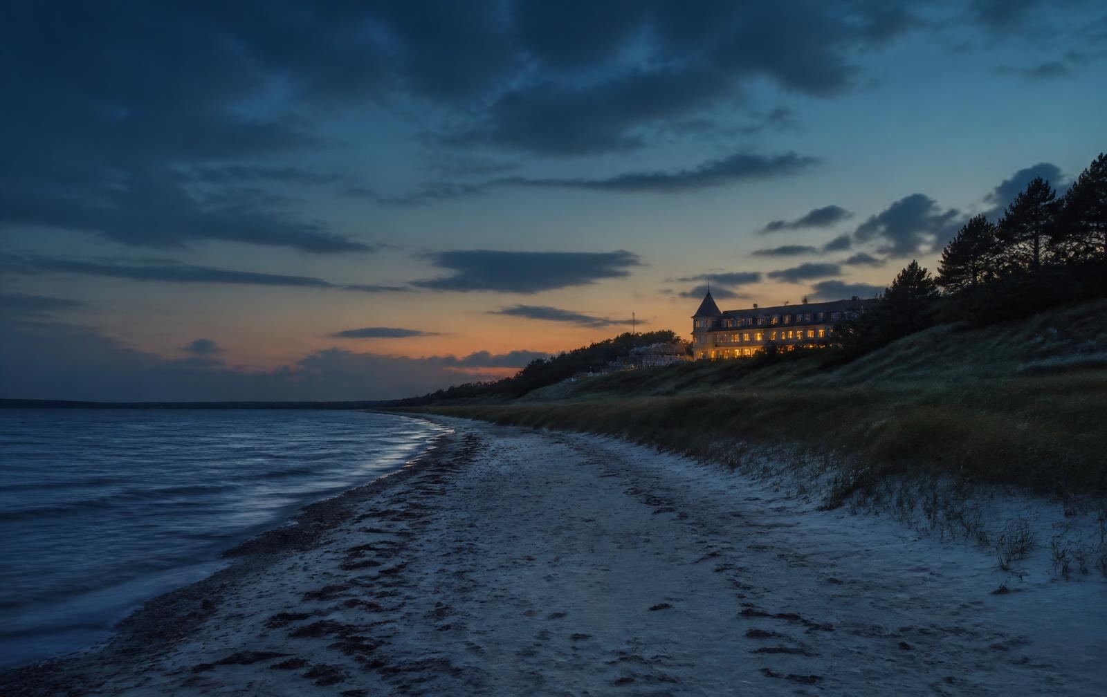Danish coastal evening with pale sand beach meeting calm dark sea, dune grass and pine, and warm window light from a coastal hotel above the shore