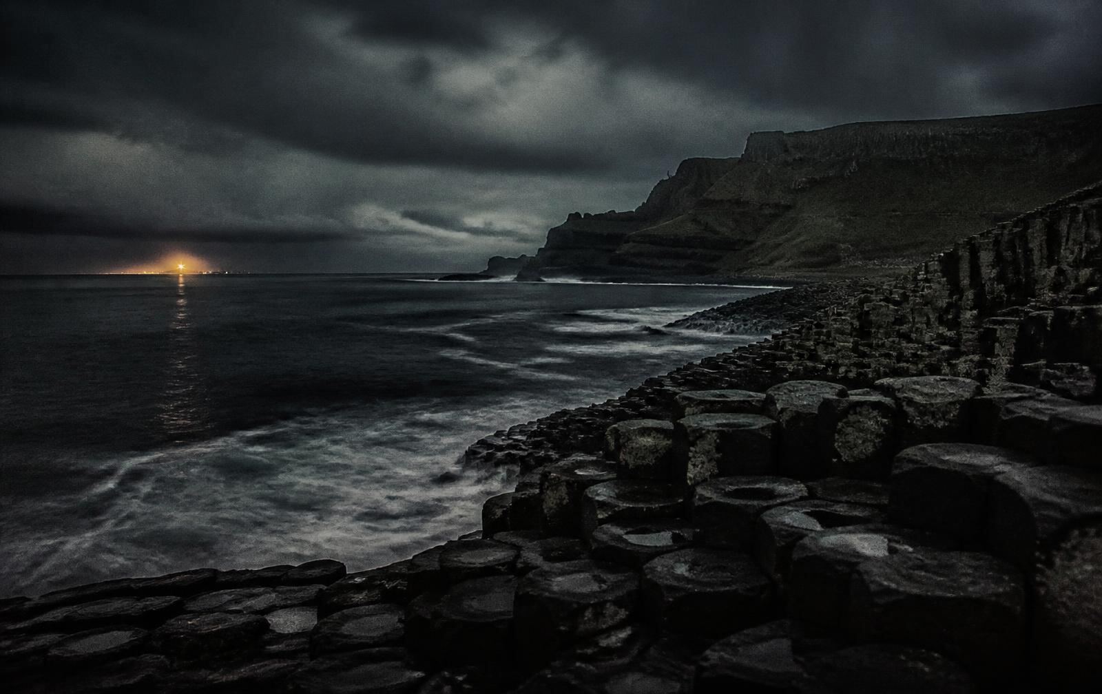 Dark basalt coastline of Northern Ireland at night under low cloud with a distant harbour light