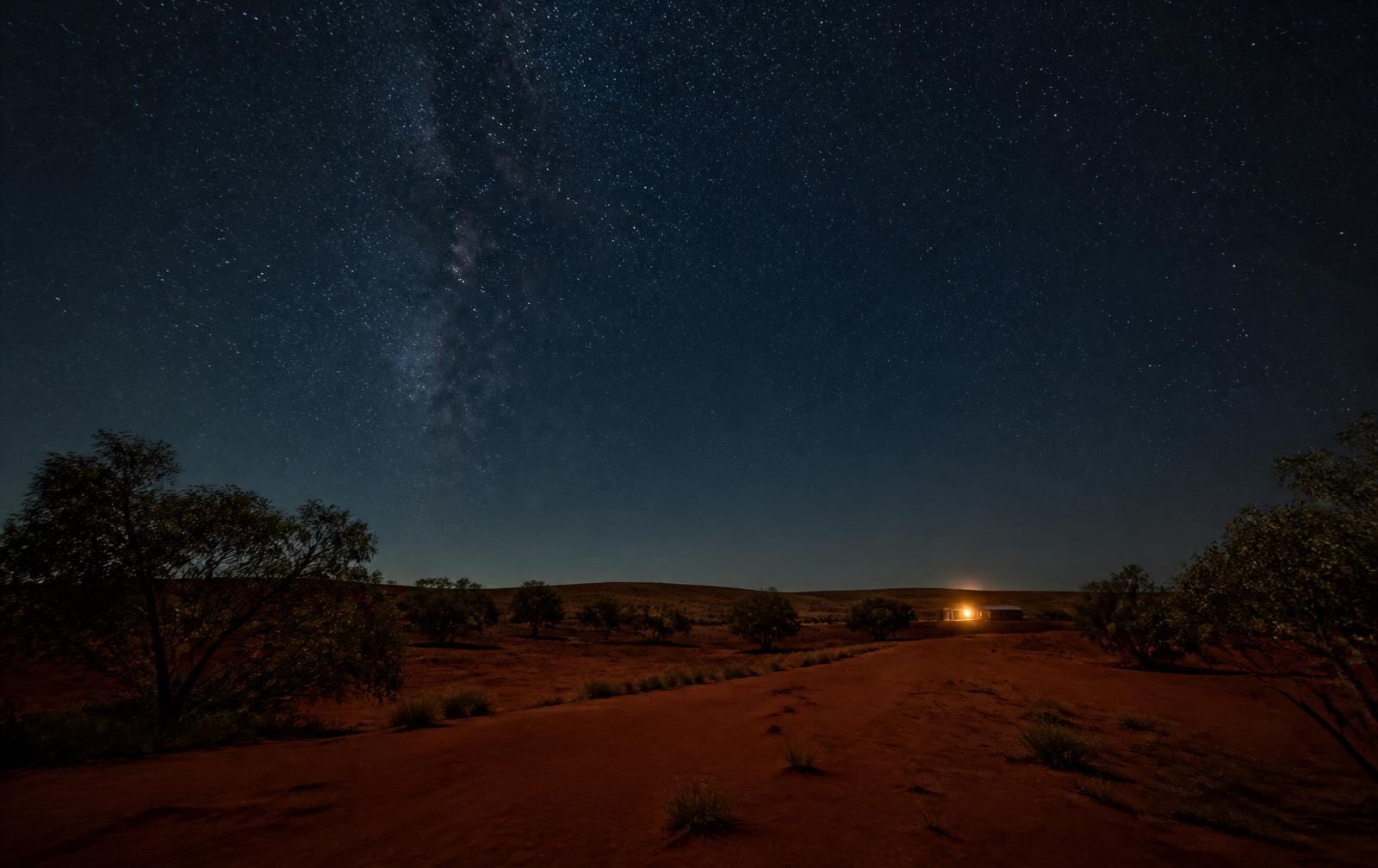 Remote red desert country at night under a vast star field with one distant warm light from a small building