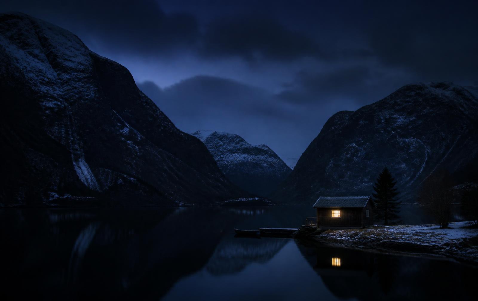 A dark Norwegian landscape at blue hour, snow-covered mountains above still black water with one warmly lit wooden building on the shore