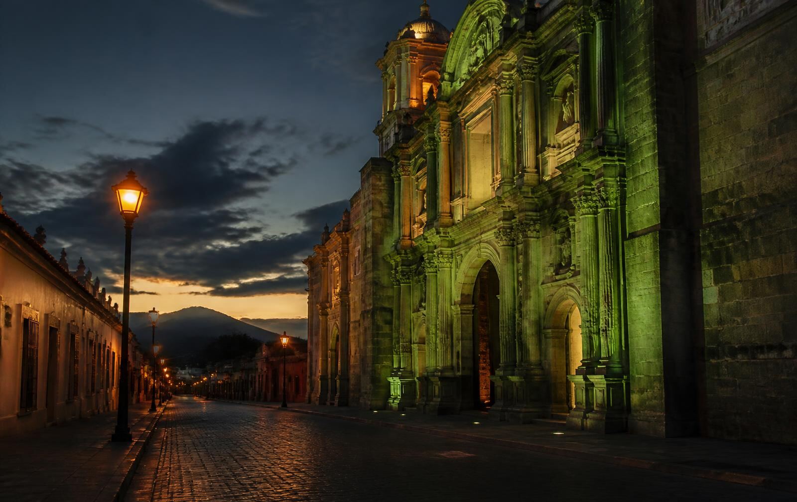 Oaxaca City's green stone church facade lit at dusk above a quiet cobbled street with lanterns