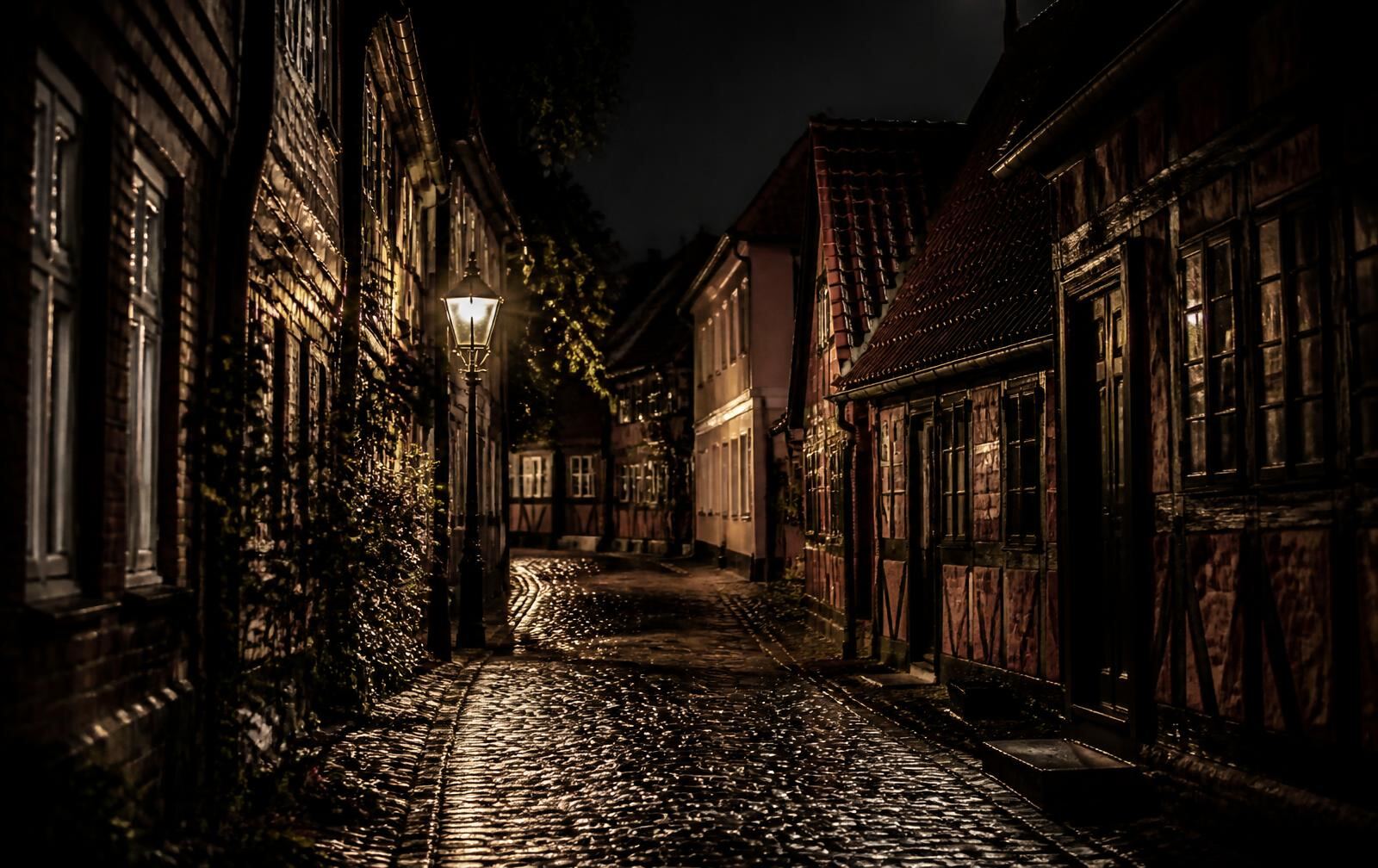 Narrow cobbled Danish street at night with half-timbered houses, a single lit lantern and wet stones reflecting warm amber light