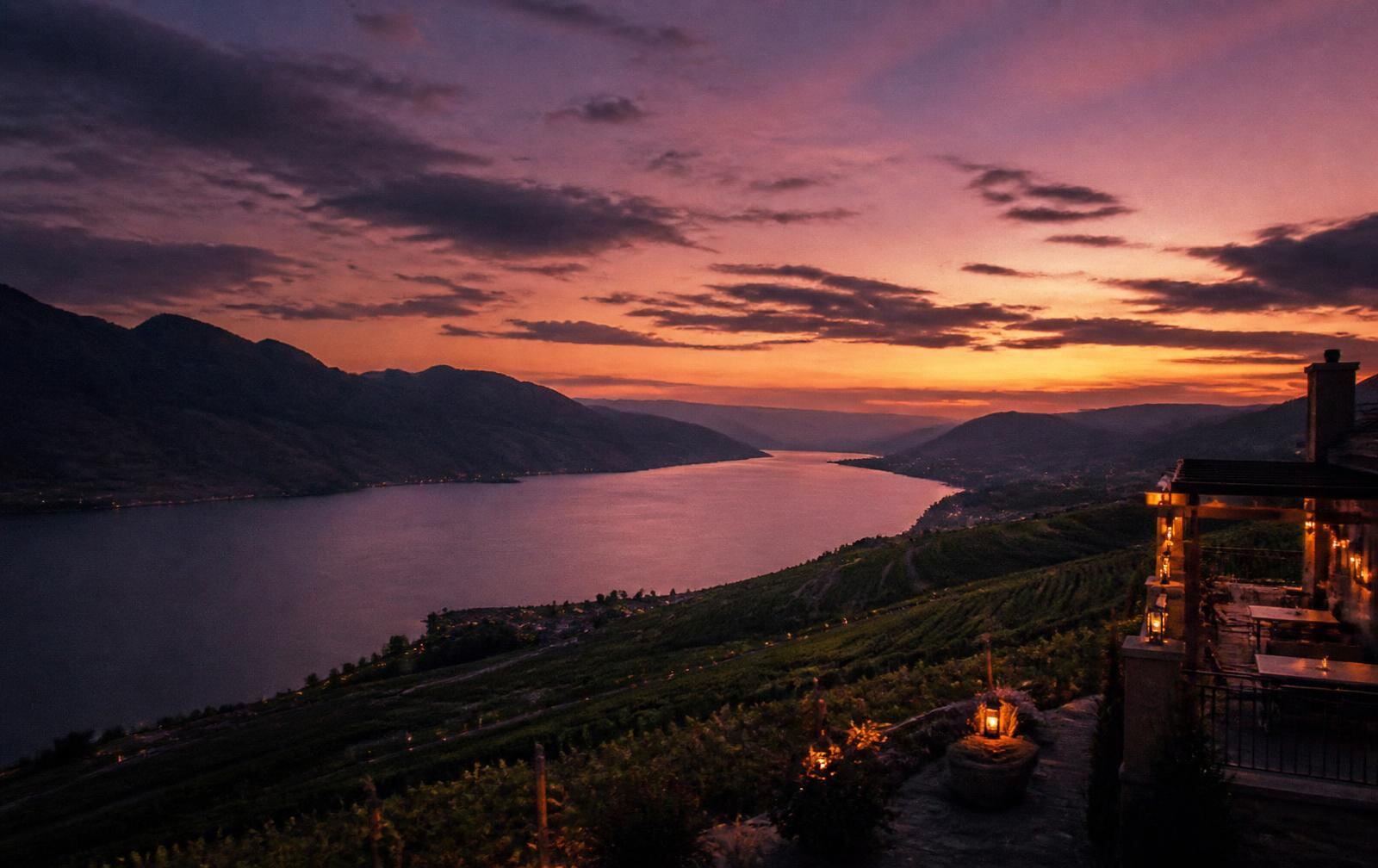 Okanagan Valley at dusk with a long dark lake below vineyard hillsides and a lit terrace in the foreground