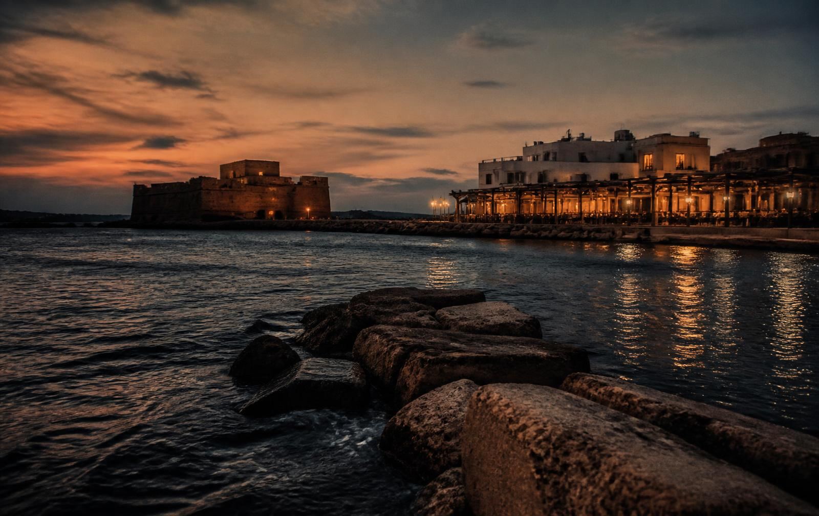 Paphos harbour at dusk with the stone fort silhouette and warm waterfront lights on dark water