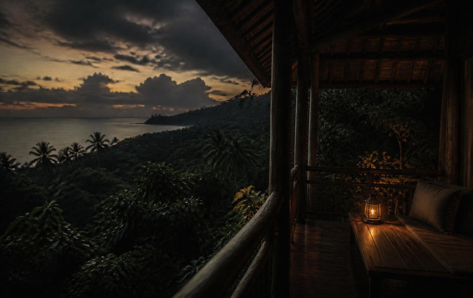 Tropical dusk over dark rainforest meeting a calm sea, seen from a timber lodge verandah with a single lantern