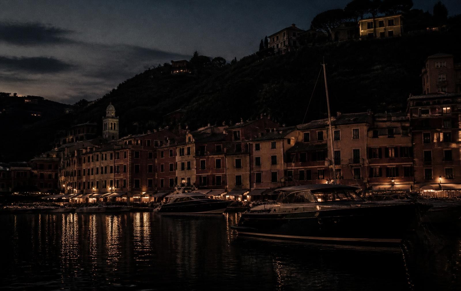 Portofino harbour at dusk with pastel facades in shadow and moored yachts lit on dark water