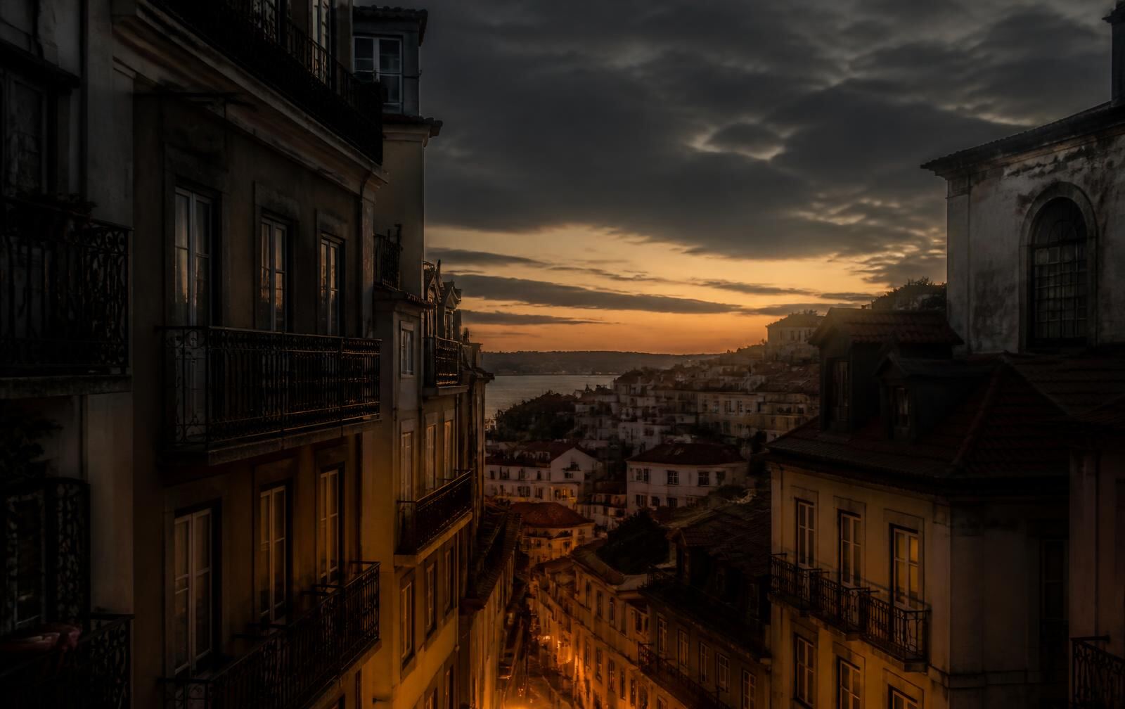 A warm stone hillside city at dusk above a wide river, with lamplit balconies in shadow