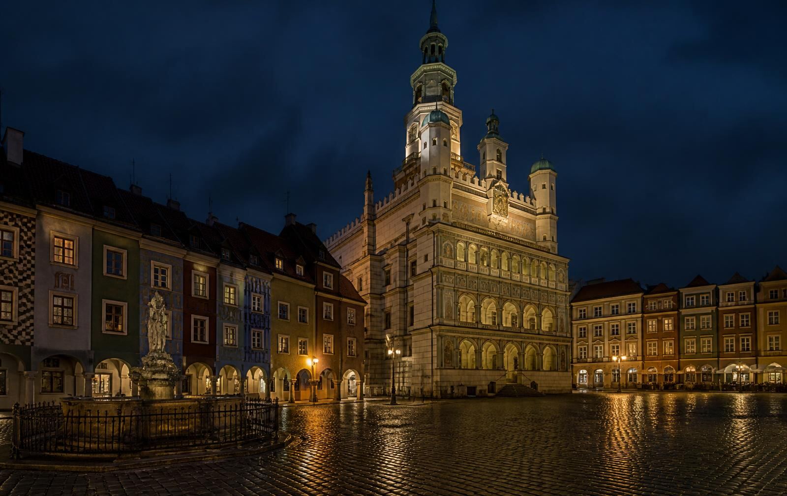 Poznań old market square at night, ornate renaissance town hall and pastel townhouses under lamplight, wet cobbles and empty arcades