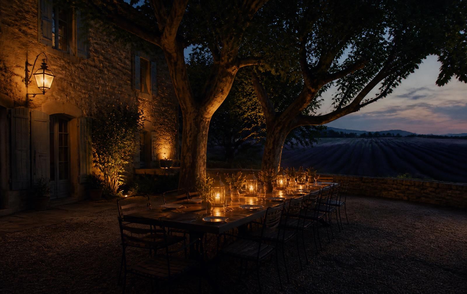 A lantern-lit stone courtyard table in Provence at dusk beneath plane trees with fields beyond