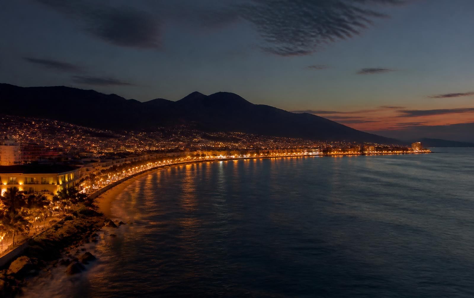 Puerto Vallarta bay at dusk, warm lights along the curving malecón with mountain silhouettes behind