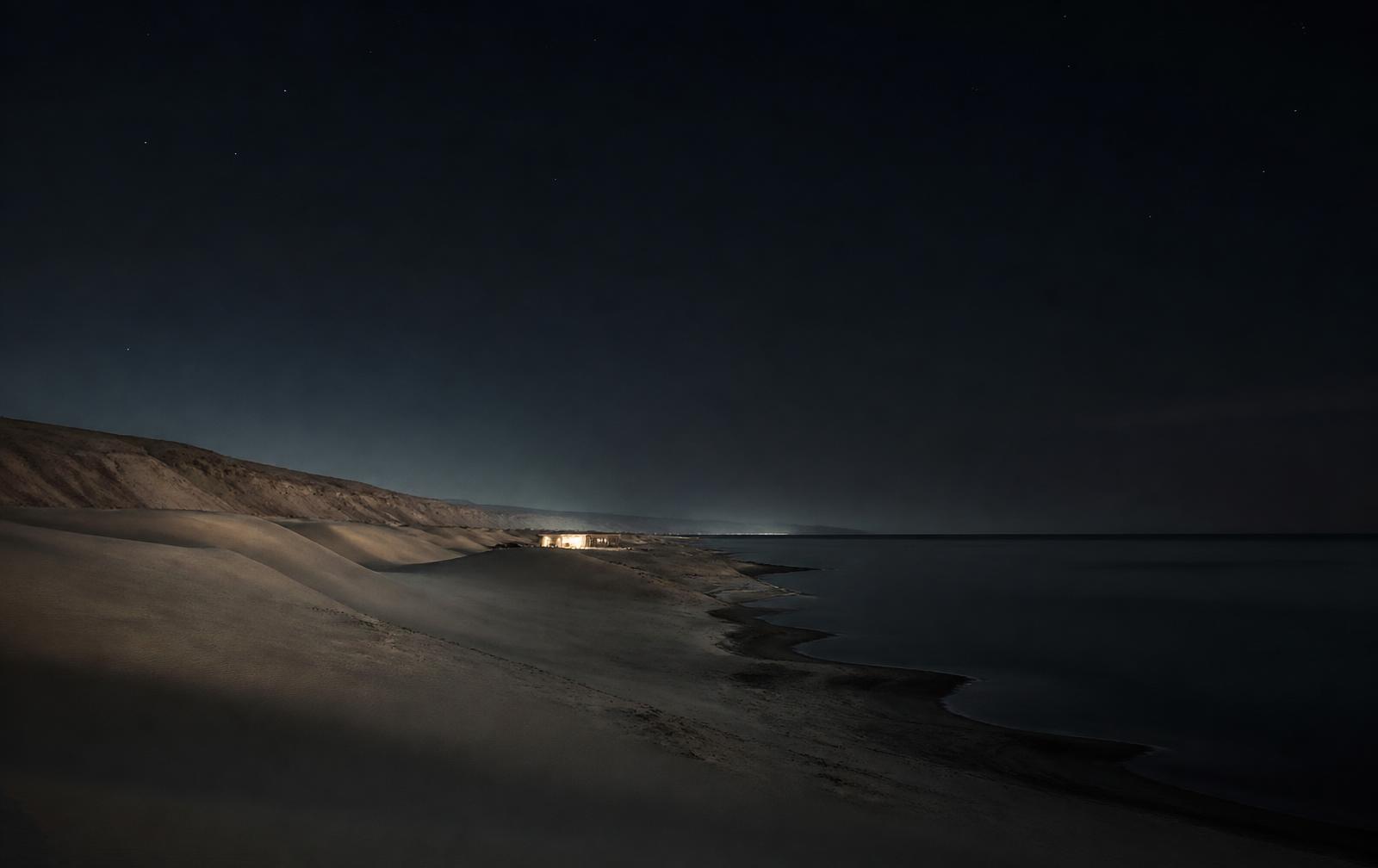 A quiet desert coastline at night where pale dunes meet very still dark sea, one faint warm light from a distant low building