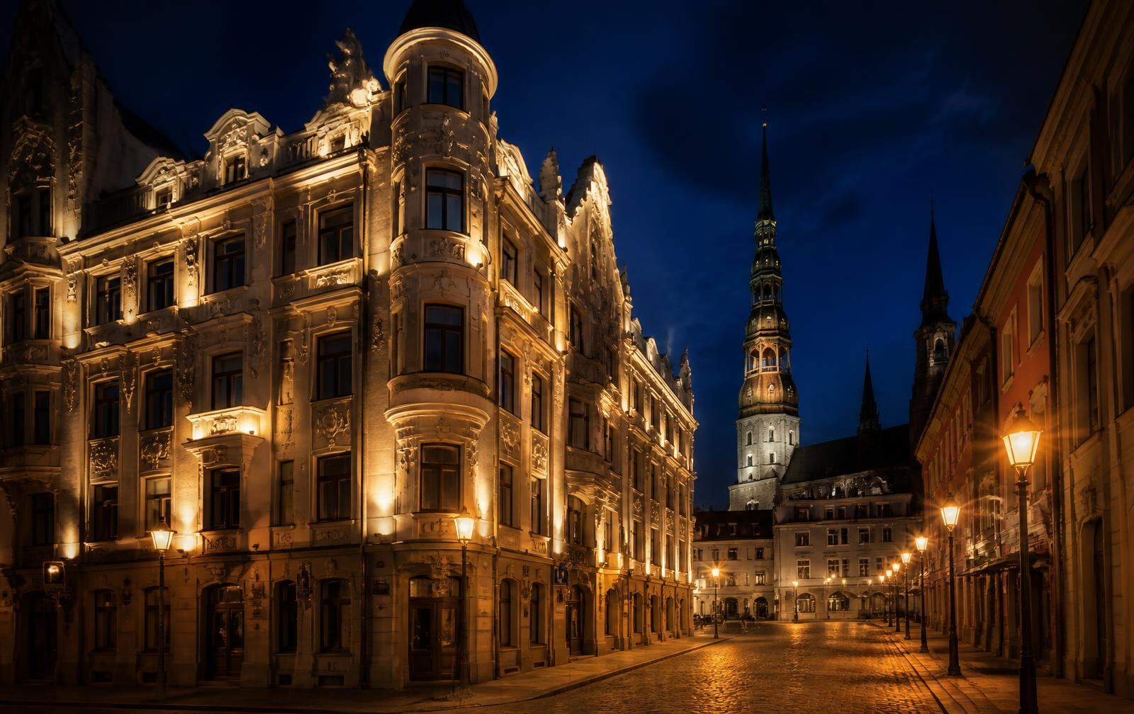 Riga's old town at night — an ornate floodlit Art Nouveau corner building, cobblestones and church spires under a deep blue sky