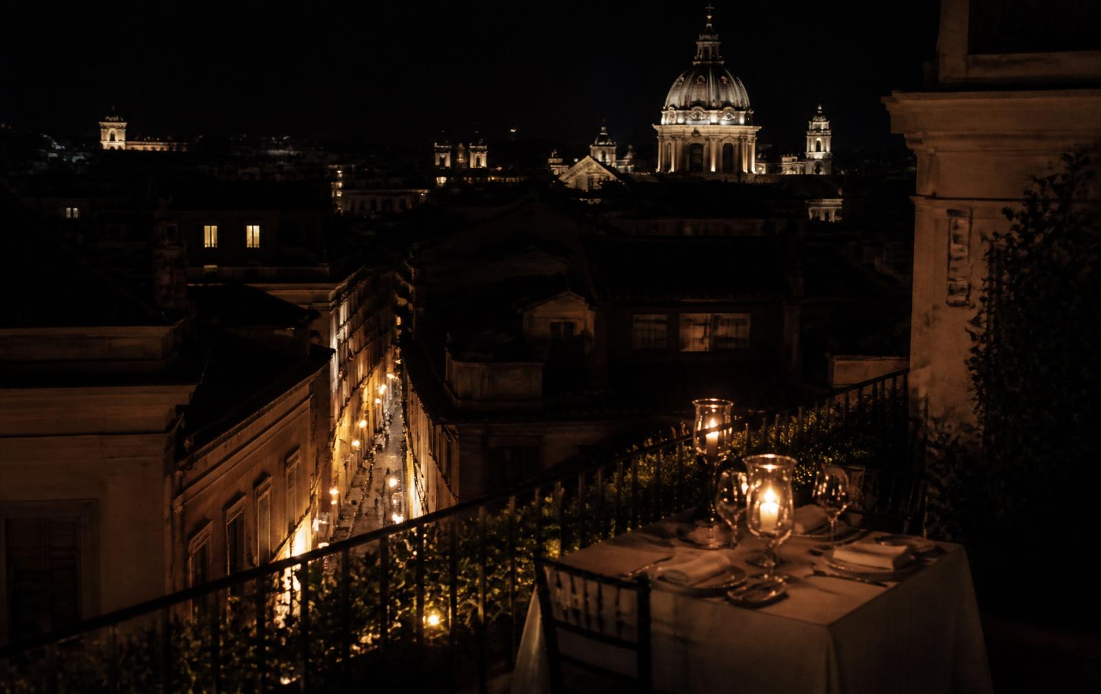 A candlelit rooftop table in Rome at night overlooking lamplit streets and a distant baroque dome
