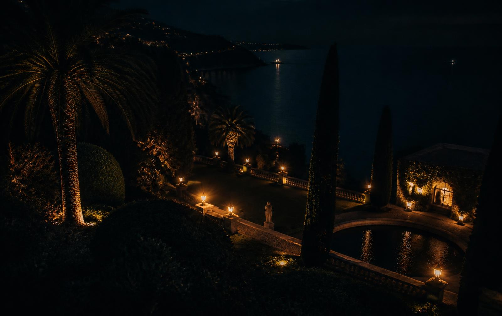 A lamplit villa garden on the Cap-Ferrat peninsula at night with cypresses above a dark sea