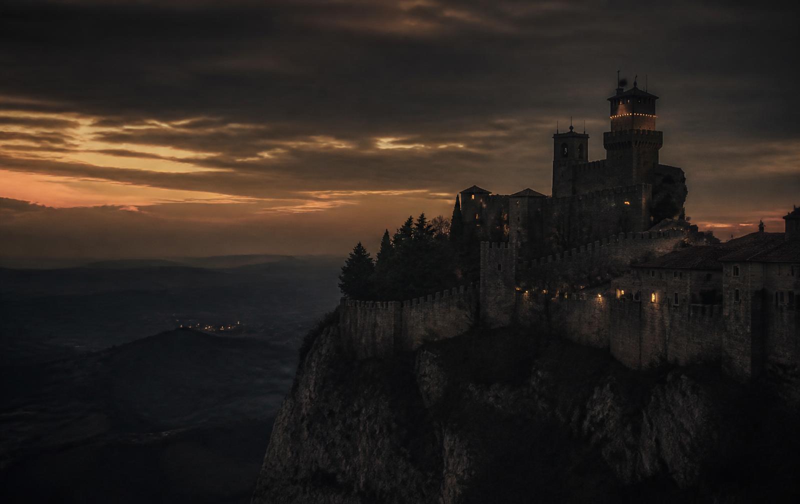 The fortified stone town of San Marino high on Monte Titano at dusk, with warm lights along the walls and a dark valley below
