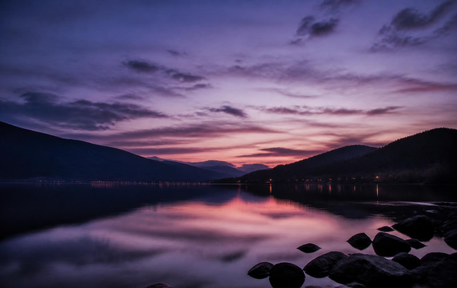 Calm freshwater lake at dusk ringed by low dark hills, with violet light on still water and a few distant shore lights