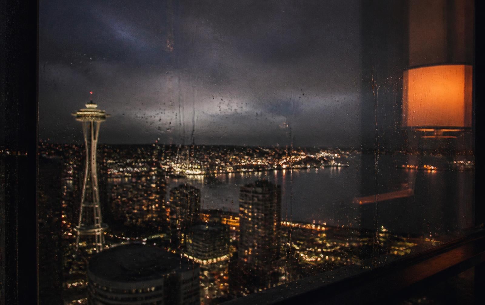 Rain-streaked window in a dark Seattle apartment with the skyline blurred behind it at night