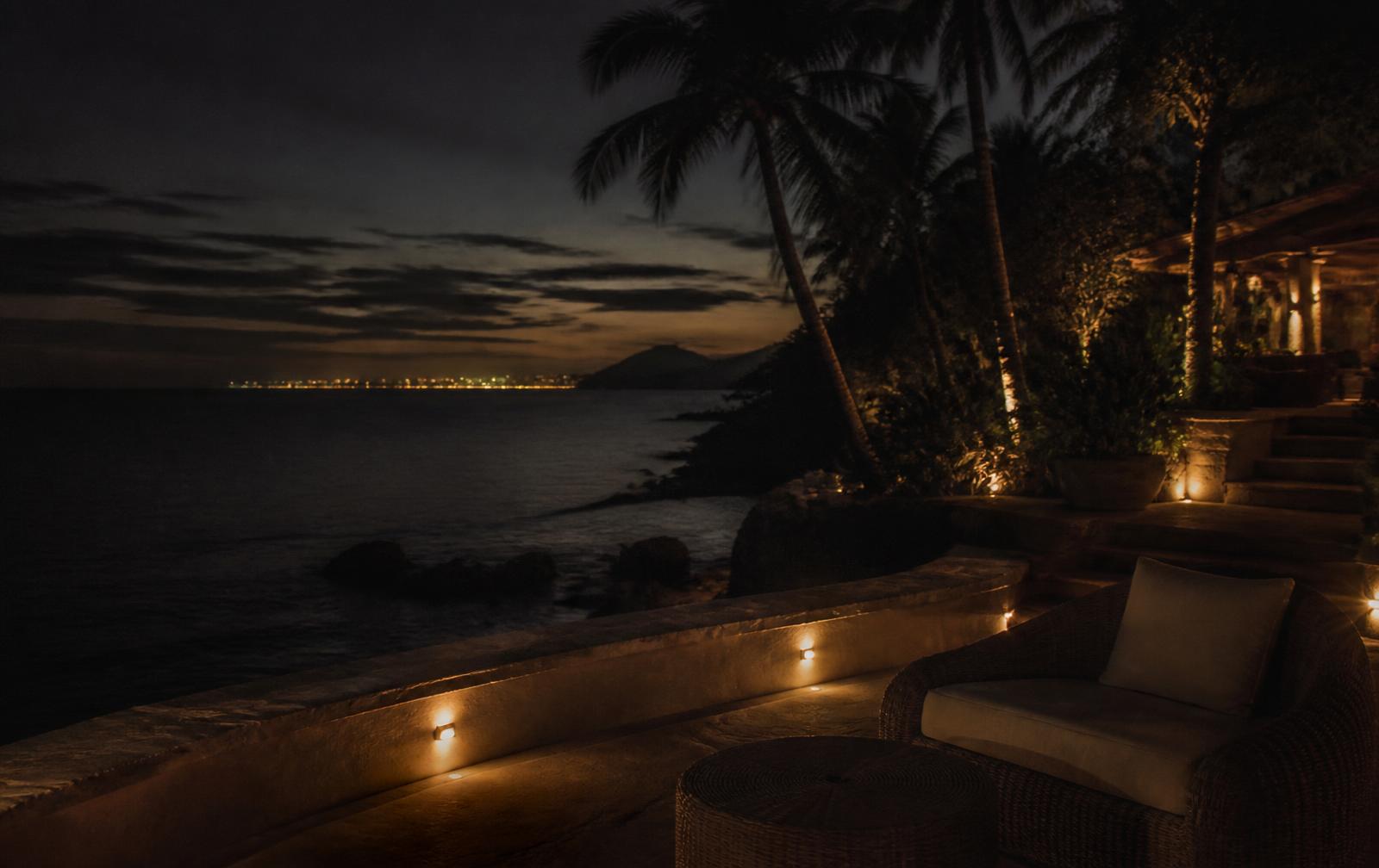 A warmly lit resort-style terrace among palms above a dark tropical sea at dusk, with faint city lights on the horizon
