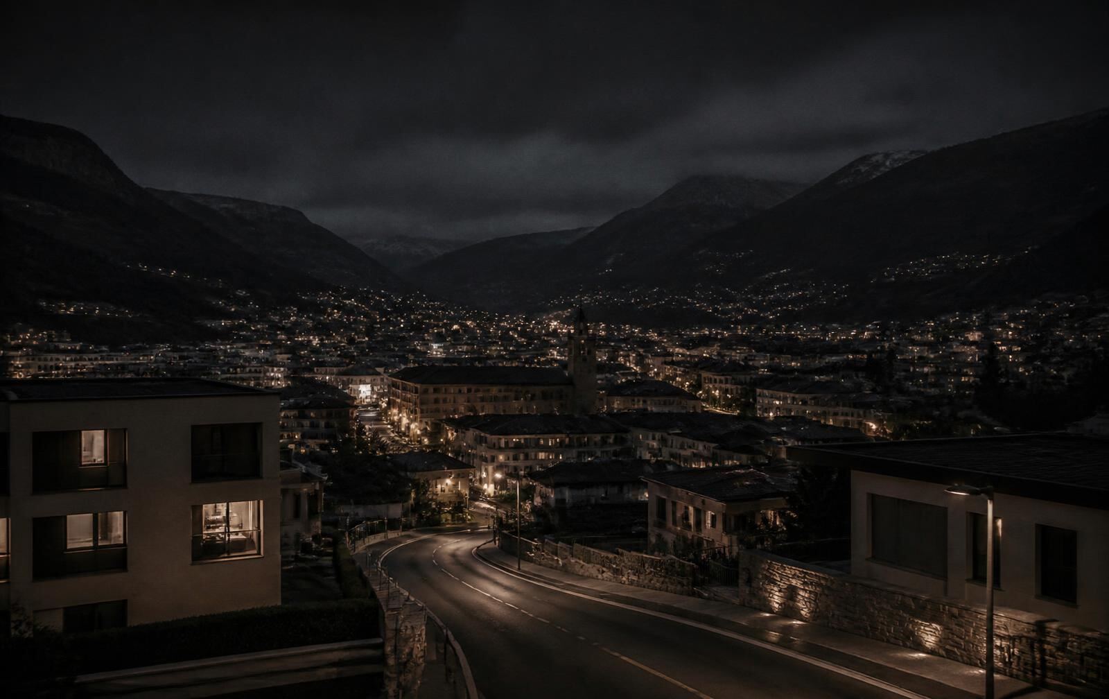 A modern low-rise town at night in a valley, lit windows and an empty curving road with dark hills behind