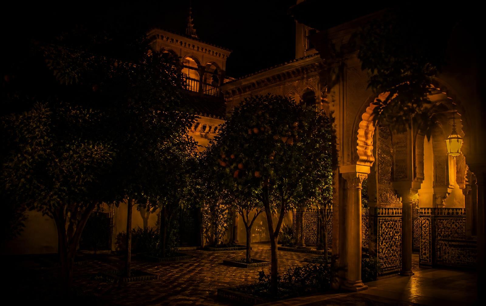 An Andalusian courtyard in Seville at night with orange trees and tiled arches under warm gold lanterns