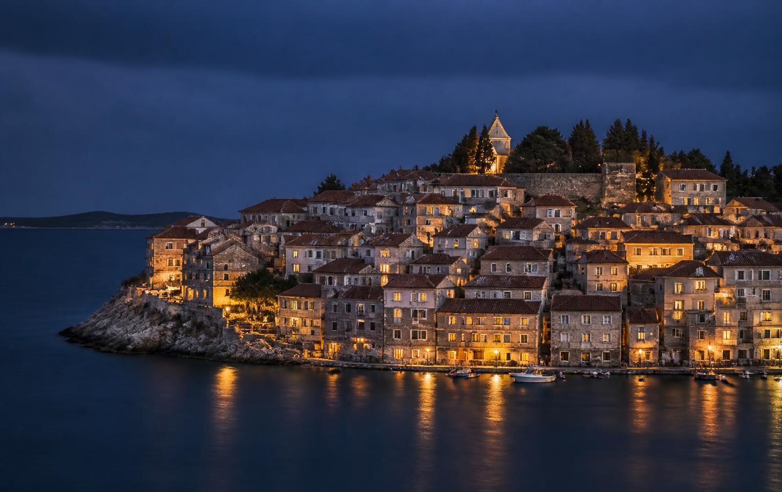 A stone Dalmatian peninsula town at dusk — honey-coloured houses above dark water under a deep blue sky