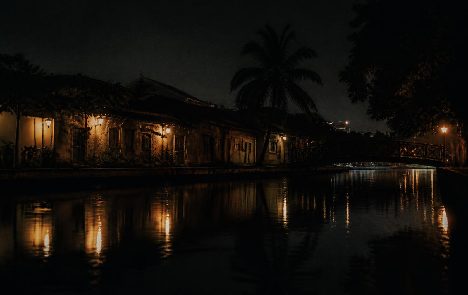 A dark riverside at night with warm lamplight reflected on still water, low old buildings and a footbridge in shadow