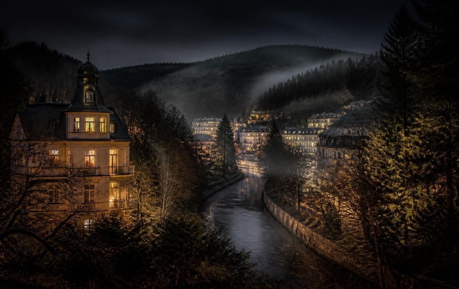 A small wooded spa town in a misty valley at night with old villas lit warm gold beside a stream