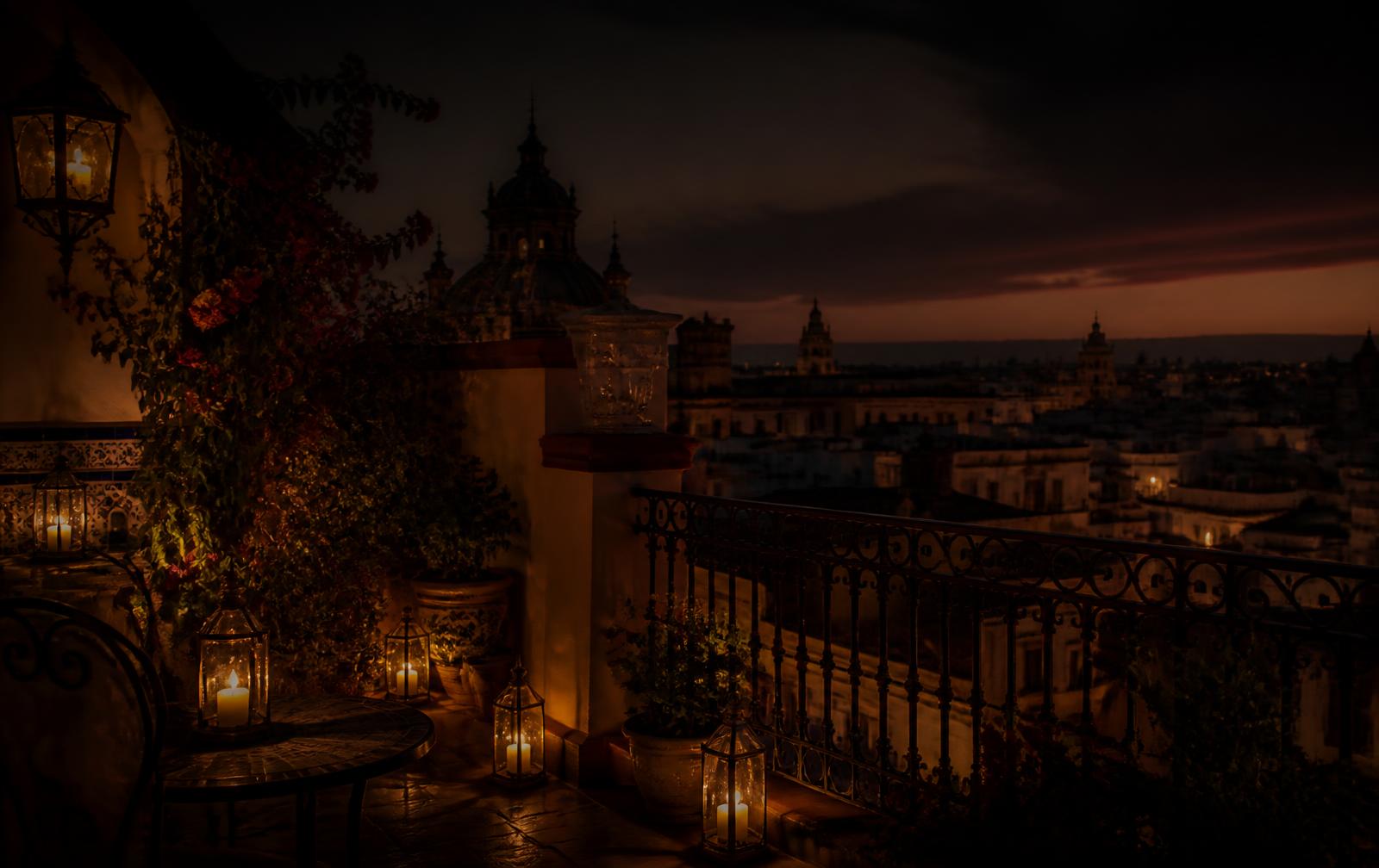 A candlelit Spanish terrace at dusk with lanterns and warm gold light above distant city rooftops