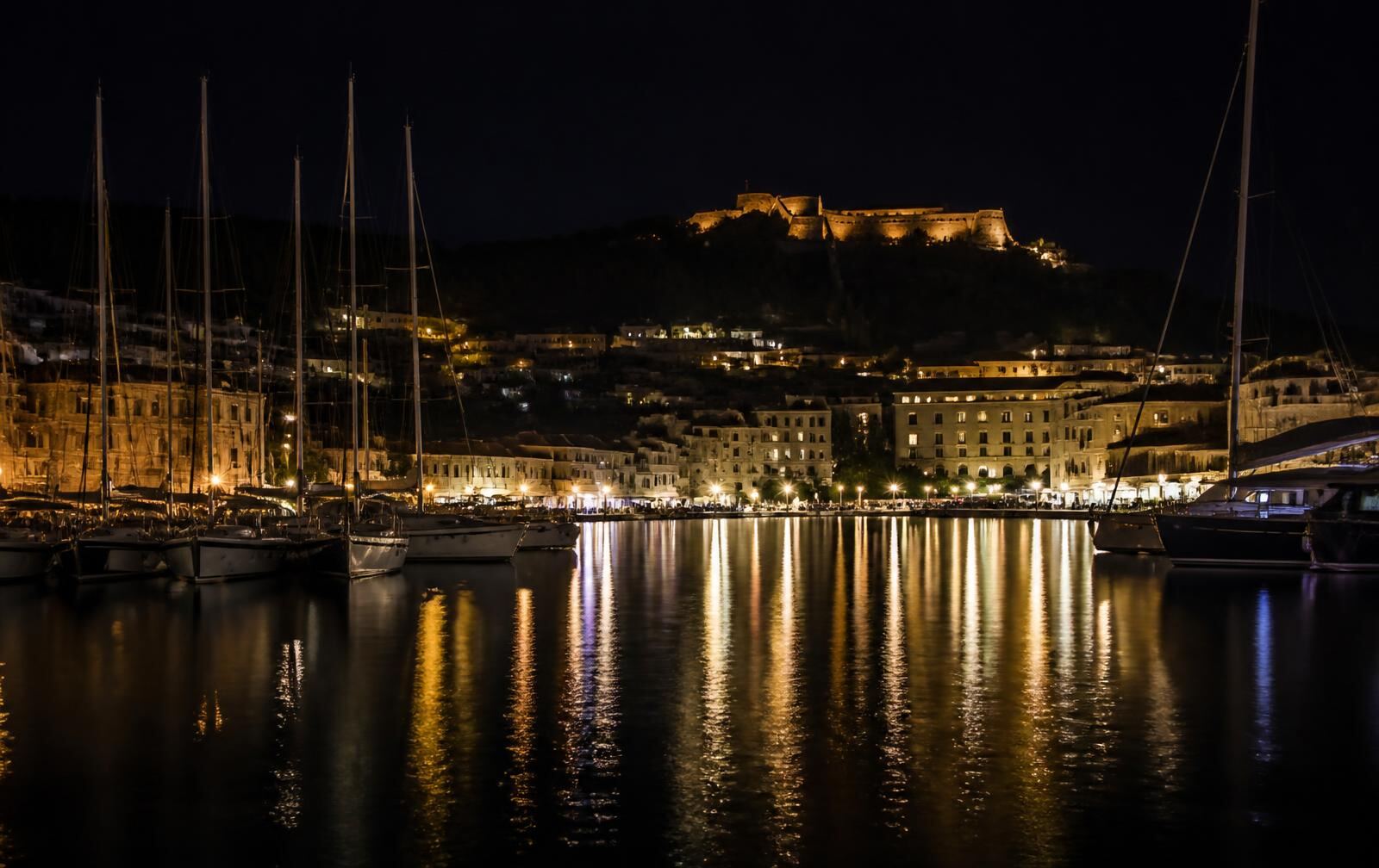 Hvar harbour at night — sailboats at rest, waterfront lights reflected in still water, a floodlit fortress above