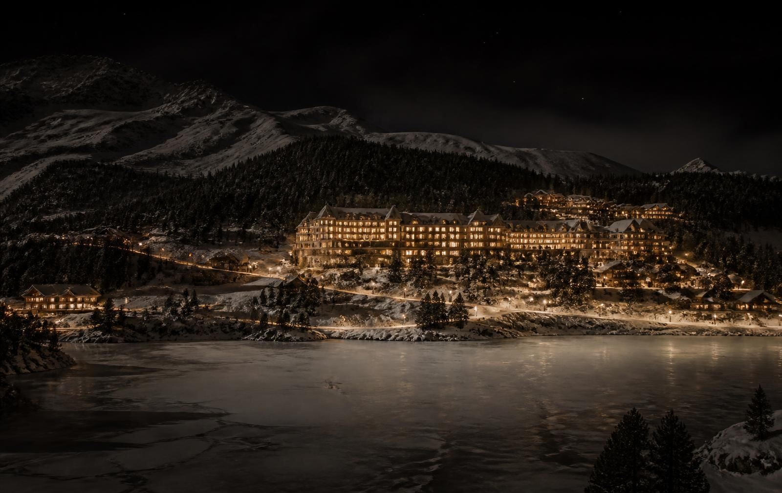 An alpine resort village above a frozen lake at night with warm gold hotel windows and snow-covered slopes