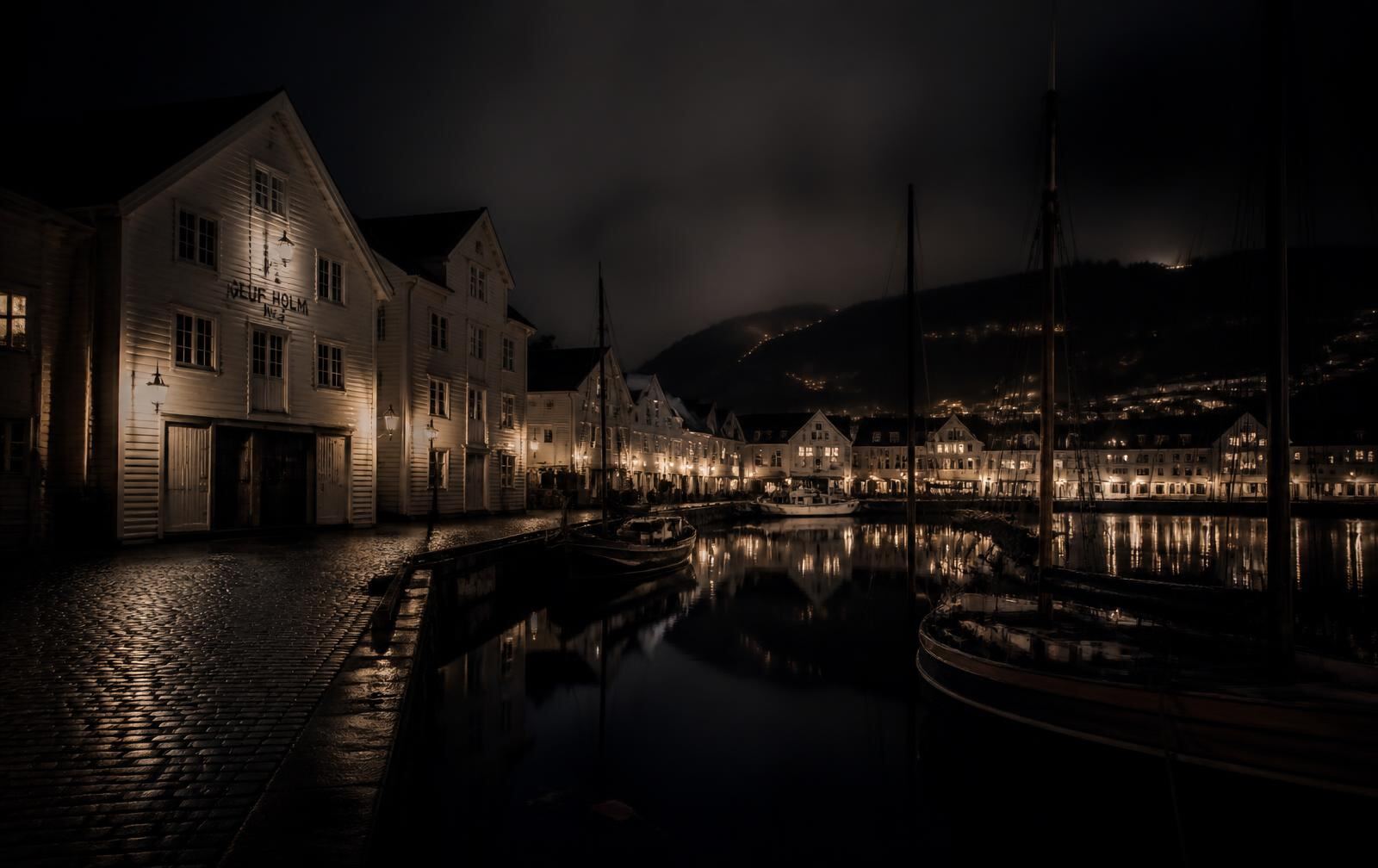 A Norwegian coastal city harbour at night, white wooden warehouse buildings lit by warm lamps, moored boats in silhouette on black water