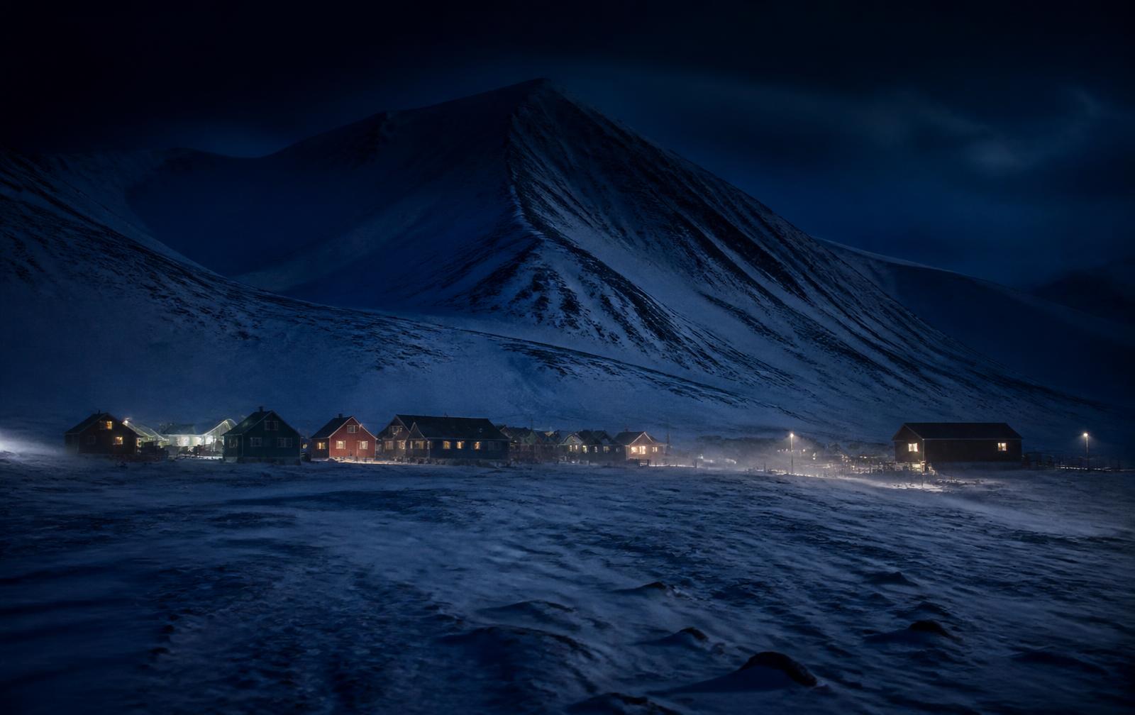 A small high-Arctic settlement during polar night, coloured wooden buildings with warm windows on a snow plain beneath dark glaciated mountains