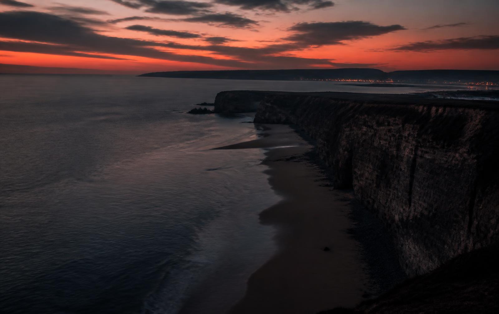 Welsh coastal cliffs at dusk above an empty beach with distant town lights along the bay