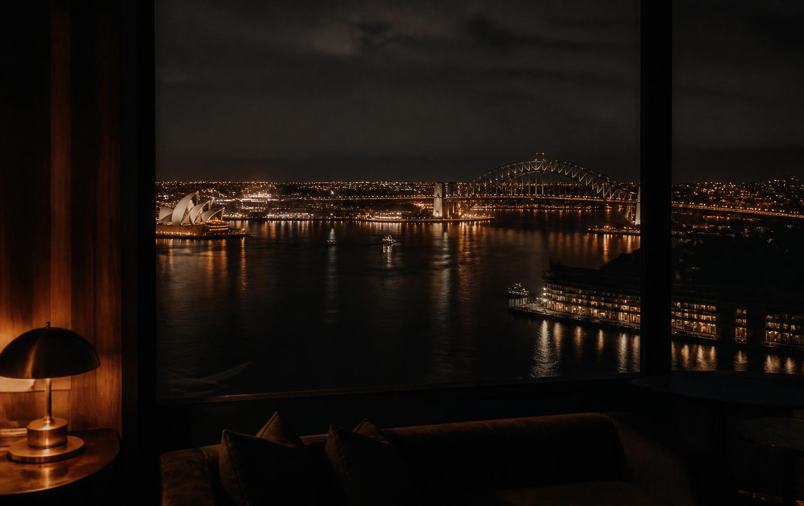 Sydney harbour at night seen from a dark timber hotel interior with a low brass lamp and gold light on the water