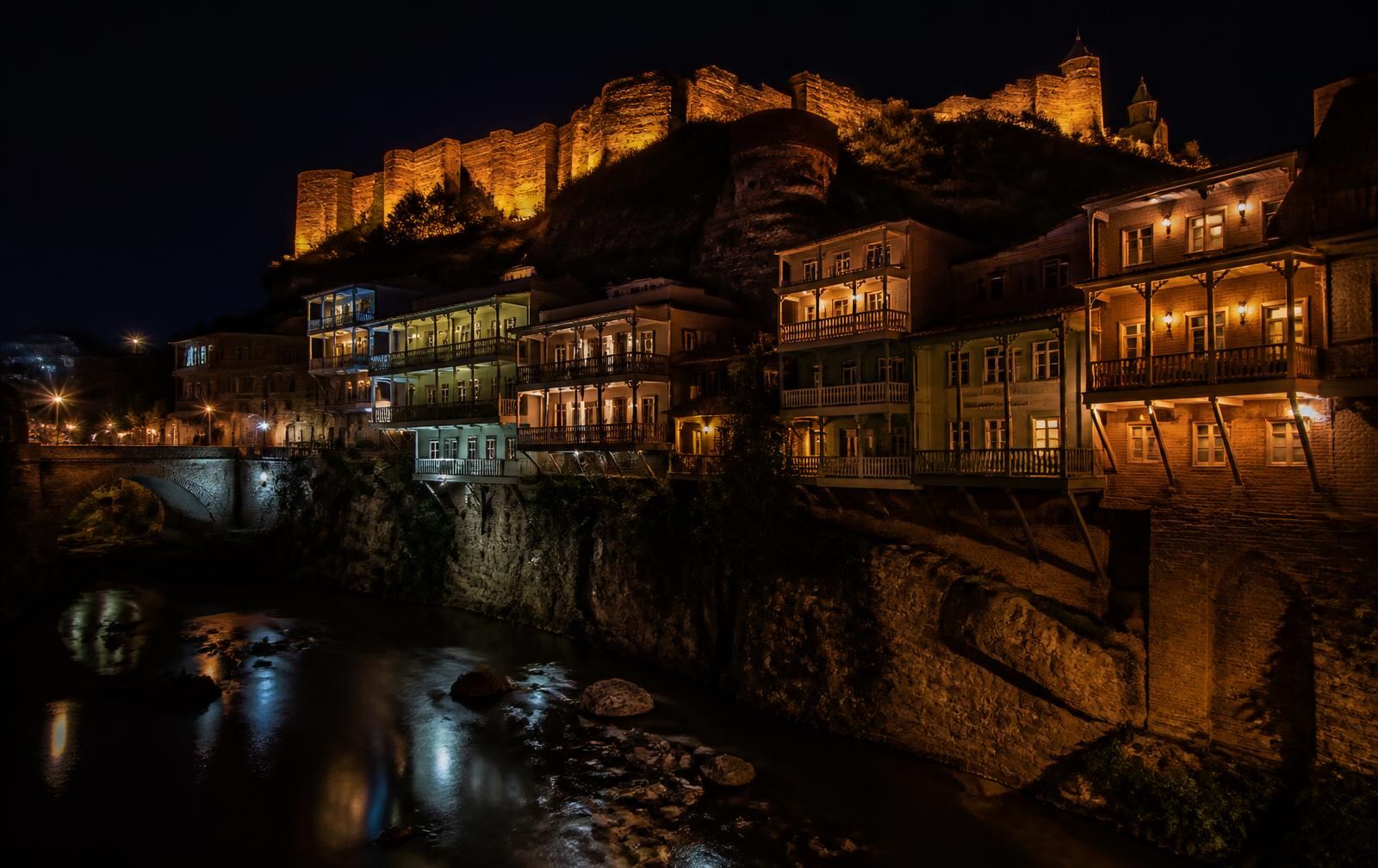 Tbilisi old town at night, warm-lit wooden balconies above the river gorge with fortress walls glowing on the hill