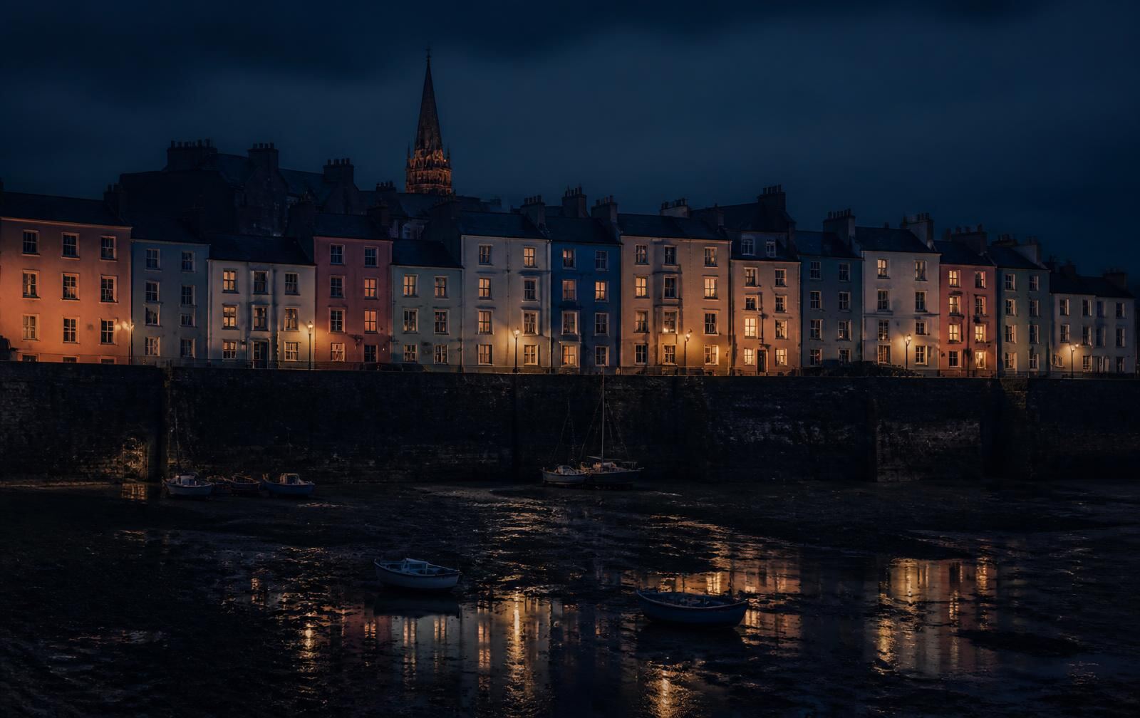 Pastel Welsh harbour houses lit at night above a dark tidal harbour with moored boats
