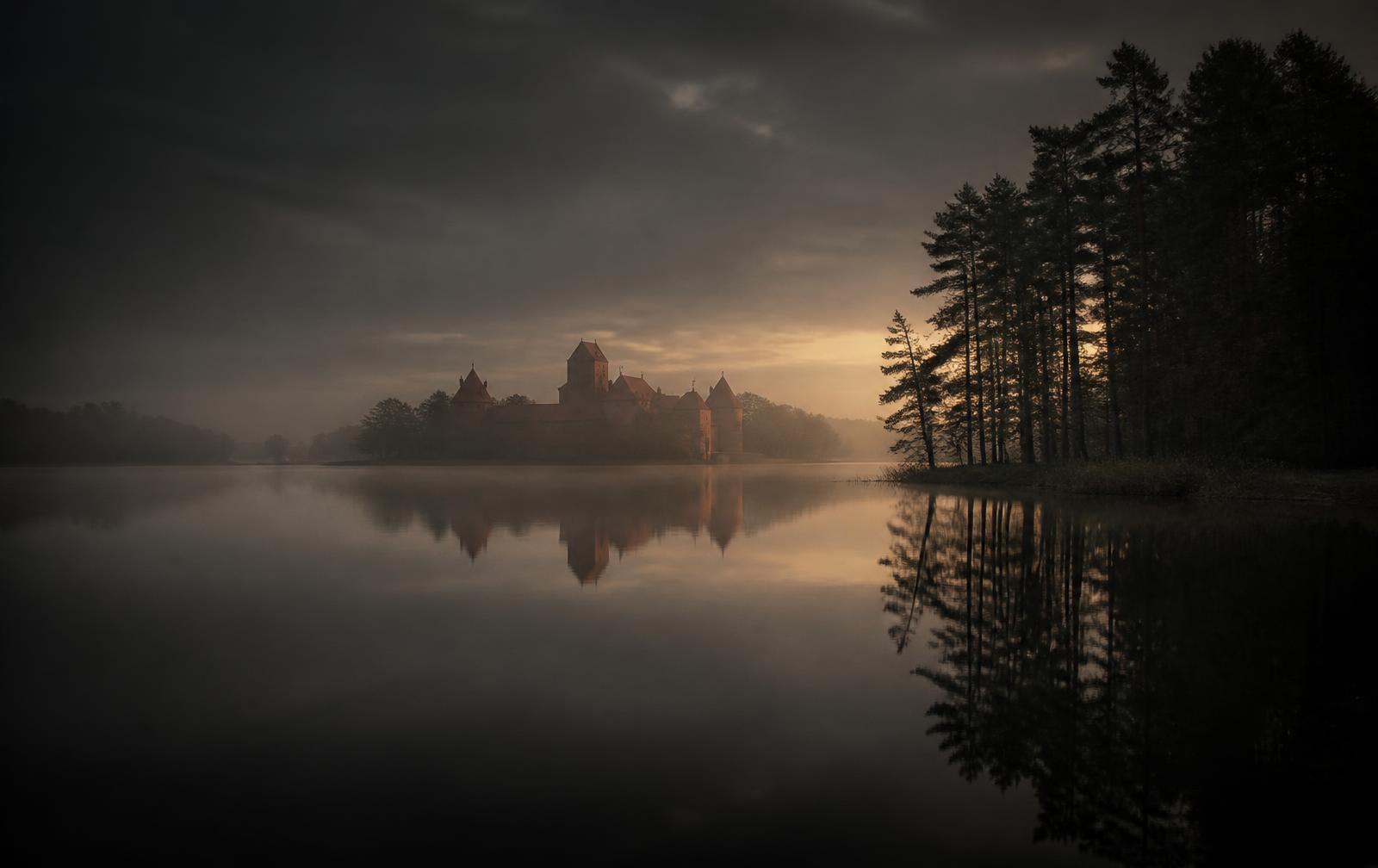 Still lake at Trakai in the evening mist, dark mirrored water, a distant red brick island castle silhouette and pine trees along the shore