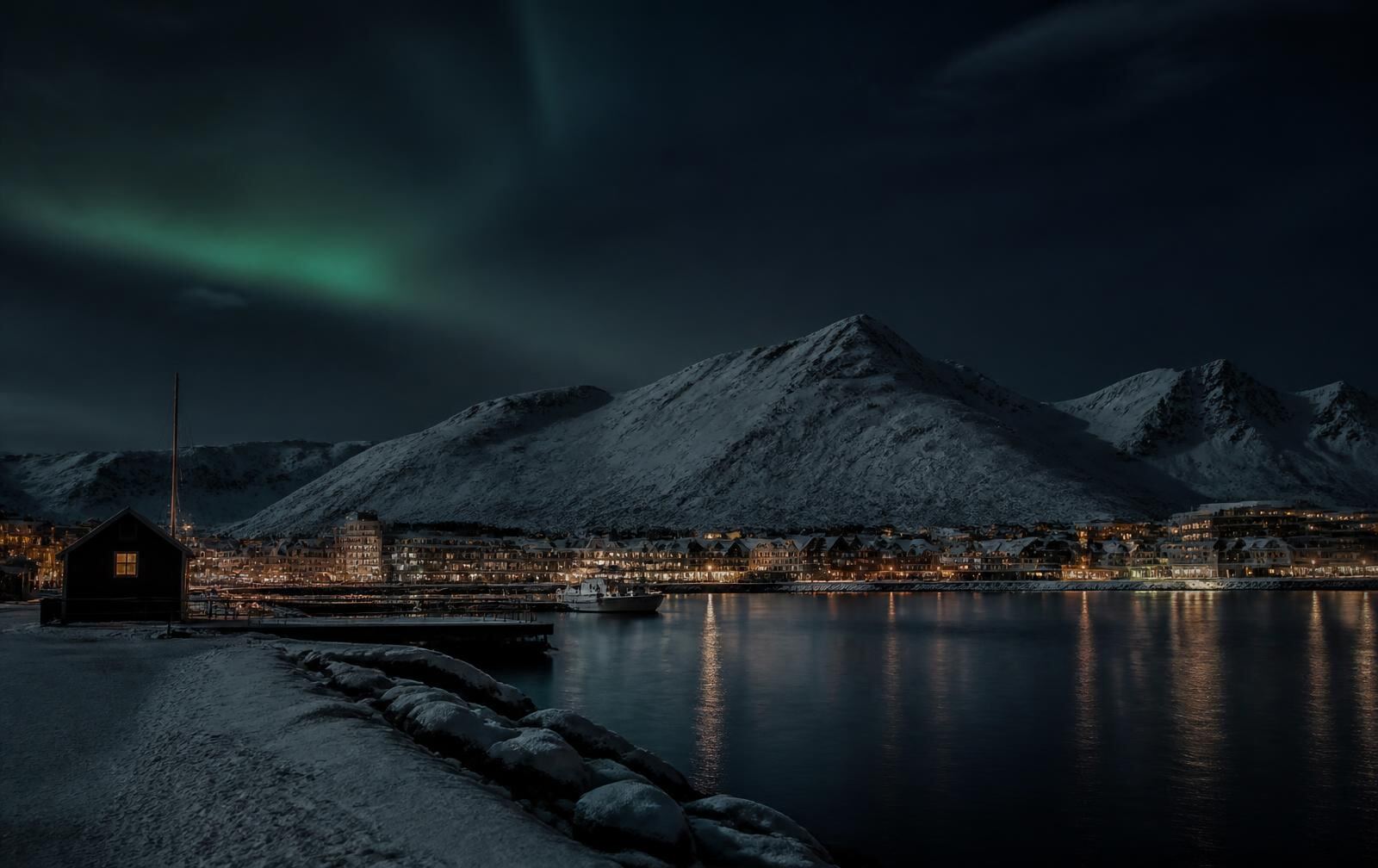 An Arctic Norwegian city at polar night, snow-covered harbour and dark mountains across the water, warm gold windows and a faint green aurora high above
