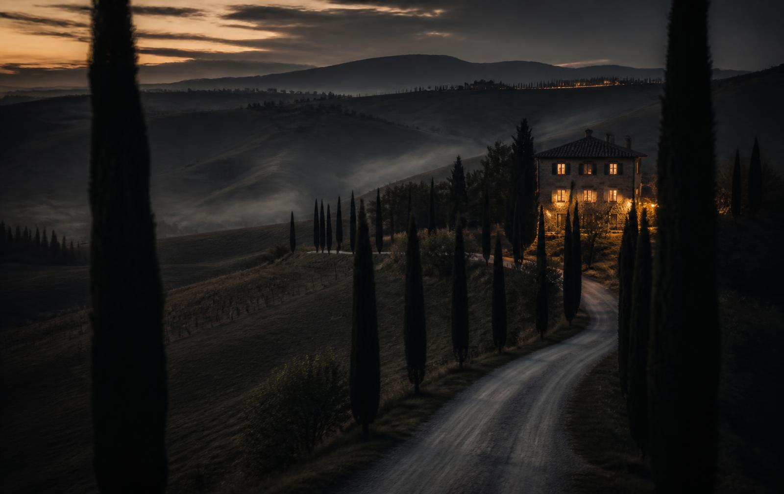 A cypress-lined road through misty Val d’Orcia hills at dusk leading to a stone villa with lit windows